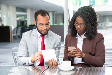 Front view of confident businesspeople using modern devices. Focused African American colleagues holding smartphone and tablet. Technology concept