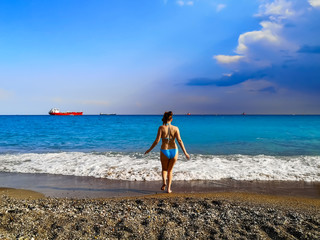 Obraz premium Seascape with many ships on the horizon and one woman on the beach. A young adult woman in a swimsuit with a slim figure enters the sea water with white foam