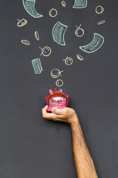Concept of saving money with a pink piggy bank held in a hand in front of a blackboard with hand drawn coins and banknotes