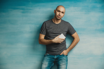 A cool bald man looks at the camera and holds out a white card, stands on a blue background, his second hand in his pocket.