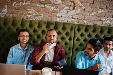 Group of four south asian men's posed at business meeting in cafe. Indians work with laptops together using various gadgets, having conversation. Hold credit card to pay.
