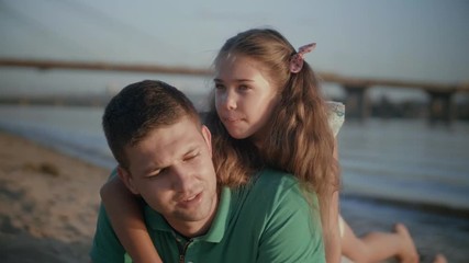 Portrait of handsome father and beautiful daughter resting on summer beach and talking while spending leisure outdoors at sunset. Charming child embracing her dad while lying on his back in nature. - Powered by Adobe