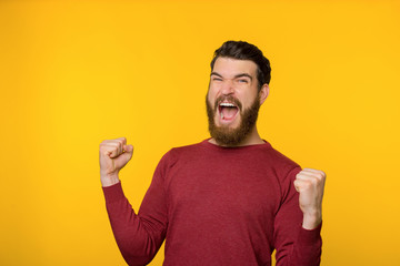 Bearded guy screaming and celebrating his triumph with rised hands, over yellow background