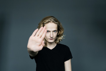 Young handsome guy with long blonde hair holds a hand in protest covering his face on a gray background.