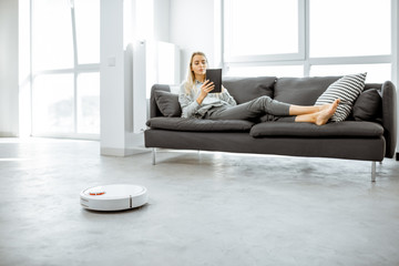 Woman relaxing on the couch while automatic vacuum cleaner doing the housework in the modern white...