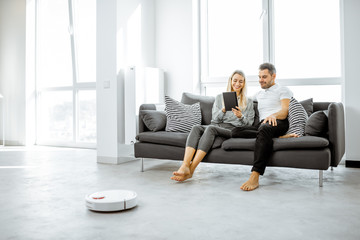 Young couple relaxing on the couch while automatic vacuum cleaner doing the housework in the modern...