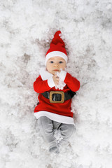 Baby in a little Santa costume lies in artificial snow, top view. Christmas card