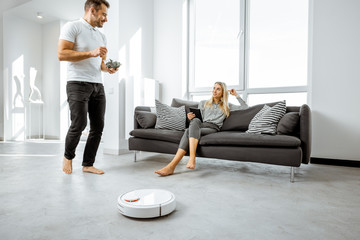 Young couple relaxing on the couch while automatic vacuum cleaner doing the housework in the modern white living room. Household robots concept