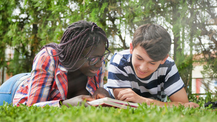 African teenager girl lying on the grass with her caucasian friend making school lesson. Happyness and Lightheartedness concept