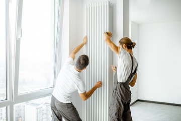 Two professional workmen in workwear installing decorative radiator in the white living room,...