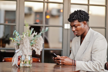 Stylish afro man in beige old school suit sitting on cafe with mobile phone. Fashionable young African male in casual jacket on bare torso.