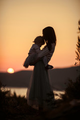 Silhouettes of mother with a baby in her arms on nature with a view of the sun setting in the water, evening at golden hour