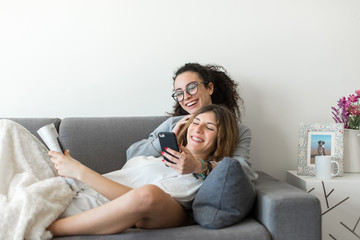 Two happy young casual women having fun using smartphone at home. Lesbian millennial couple.
