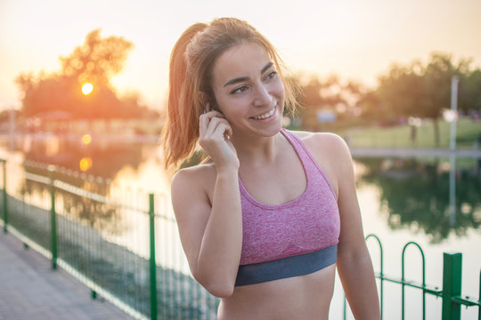 Smiling young fitness woman with airpods listening to music during her outdoor workout in the park