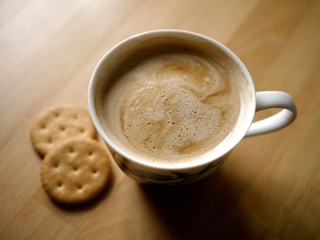 Photo of a cup of coffee with foam and two cracker cookies on a light brown wooden table. Coffee and snacks concept.