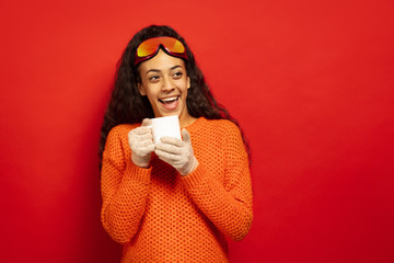 African-american young brunette woman's portrait in ski mask on red studio background. Concept of human emotions, facial expression, sales, ad, winter sport and holidays. Drinks tea, coffee.