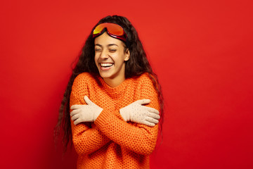 African-american young brunette woman's portrait in ski mask on red studio background. Concept of human emotions, facial expression, sales, ad, winter sport and holidays. Warming in cold, laughting.