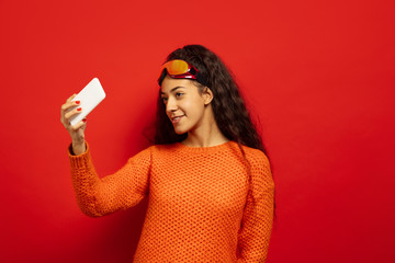 African-american young brunette woman's portrait in ski mask on red studio background. Concept of human emotions, facial expression, sales, ad, winter sport and holidays. Drinks tea, coffee.