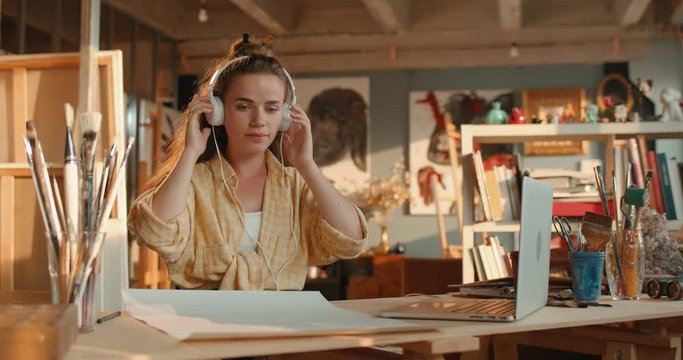 Attractive young Caucasian female artist taking on headphones and listening to the music while depicting a picture from the screen of the laptop computer while drawing with a pencil.