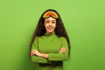 African-american young brunette woman's portrait in ski mask on green studio background. Concept of human emotions, facial expression, sales, ad, winter sport and holidays. Smiling, hands crossed.