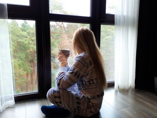  fair-skinned young girl with blond long hair in pajamas sits by the window with a gray cup and a kitten