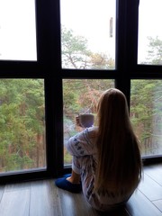  fair-skinned young girl with blond long hair in pajamas sits by the window with a gray cup and a kitten