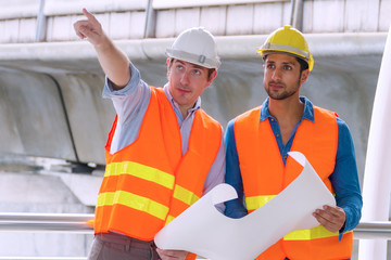 Two engineer or architect with safety helmet planning about building with blueprint. Professional technician man pointing finger while his colleagues looking at construction site.Industry work concept