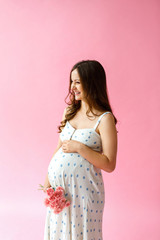 Studio shot of happy pregnant woman dressed in comfort home dress on a pink background, touching gently her belly and smiling at camera. Pregnancy healthy motherhood concept.