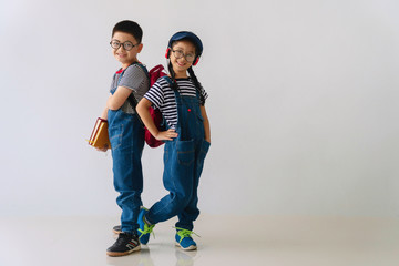 Cheerful brother and sister wear eyeglasses standing back to back together on white background. Little boy hold books in his hand and little girl smile wear earphones with hat. Back to school concept.
