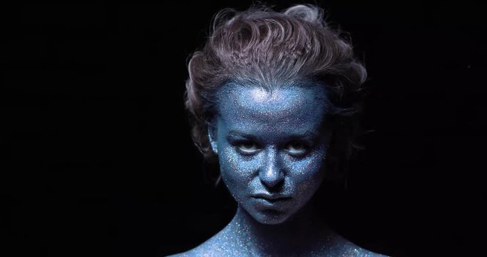 girl with a blue face in sparkles close-up against a black wall looks at the camera