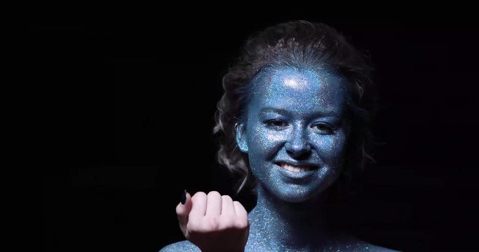 close-up of a girl with a blue face in sparkles against a black wall blows sparkles from her palm right into the camera