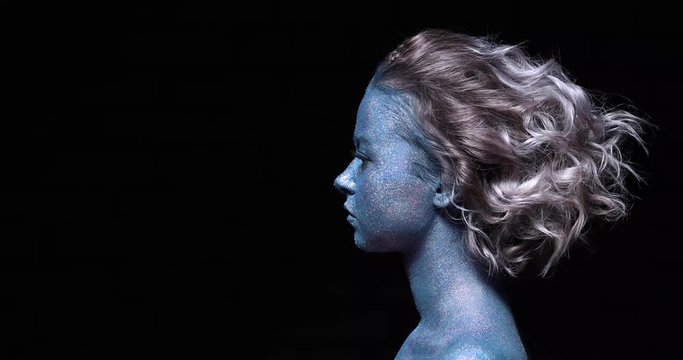 a girl with a blue face in profile in sparkles close-up against a black wall looks forward