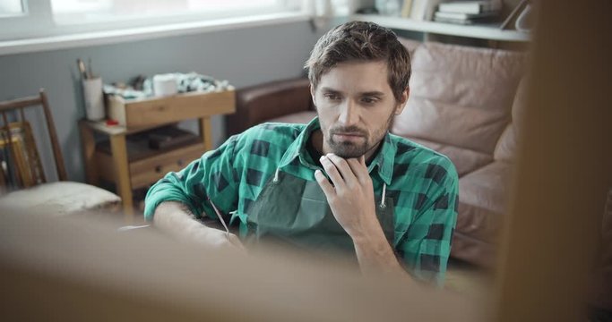 Young handsome focused male artist sitting in front on molbert, developing picture idea, Bohemia