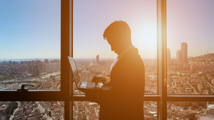 Silhouette of businessman is working on his laptop in office. He is standing near the panoramic...