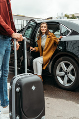 Driver holding wheeled suitcase and smiling woman sitting in taxi