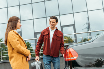 Taxi driver holding suitcase and smiling to passenger by car trunk