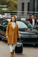 Young woman with suitcase and smiling taxi driver by car at background