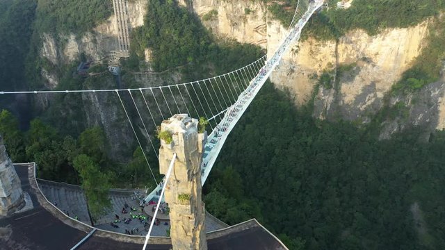 Tilting aerial view of tourists walking over glass suspension bridge, looking around 300 meters down on beautiful mountain valley, landmark travel destination in China