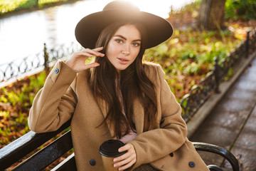 Portrait of alluring woman sitting on bench with coffee cup in autumn park