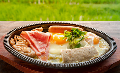 Breakfast Asian style on hot plate. KAI KATA  that made from fried egg with pepper and some meat such as chinease sausage , pork on a wooden table Amid the view of the rice fields backgroud.