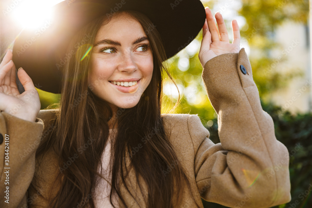 Sticker Portrait of positive woman touching her hat while walking outdoors