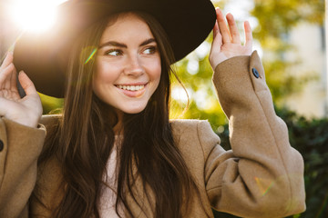 Portrait of positive woman touching her hat while walking outdoors