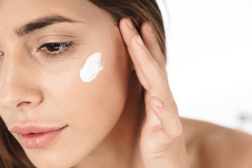 Portrait of young half-naked woman looking aside and applying facial cream