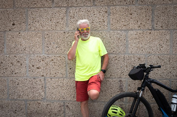Fototapeta na wymiar an old man dressed in red and yellow colors leaning against the concrete wall talking on the phone. Electric bicycle near him