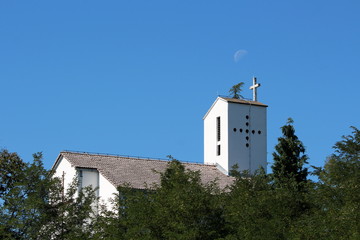 Narrow white catholic church with concrete cross on top completely surrounded with dense trees and forest vegetation on clear blue sky with barely visible moon in background on warm summer day