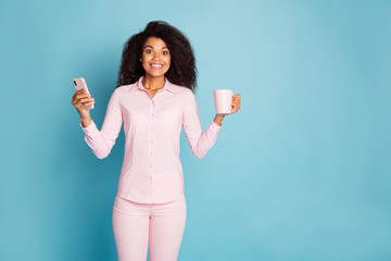 Photo of pretty wavy dark skin lady holding telephone and hot beverage mug enjoy amazing morning wear pink shirt trousers isolated blue color background