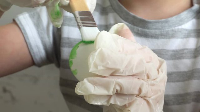 Little boy wearing protective gloves draws on an egg making an Easter drawing. She uses a brush to make different colors.