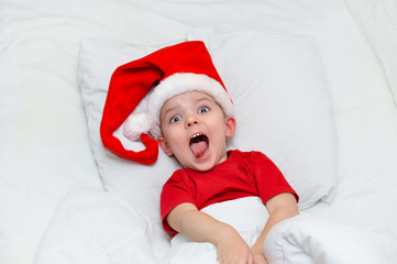 Crazy Little Santa with his mouth wide open in shock lies in his bed under a white blanket in a Santa hat.