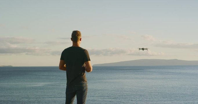 A young man flying his new quadcopter drone out over the ocean at sunset, drone pilot flying at sunset