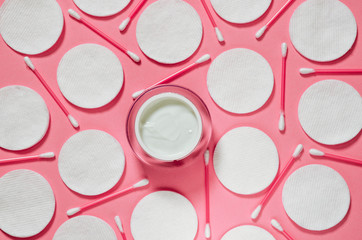 Hygiene products: white round cotton pads and plastic sticks for cleaning the ears lie on a pink background next to face cream